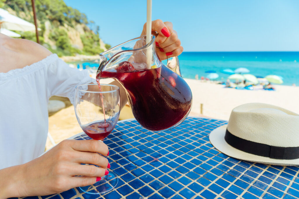 Image of a woman tourist pouring fresh red wine sangria in the glass sitting in a beach cafe (chiringuito)

