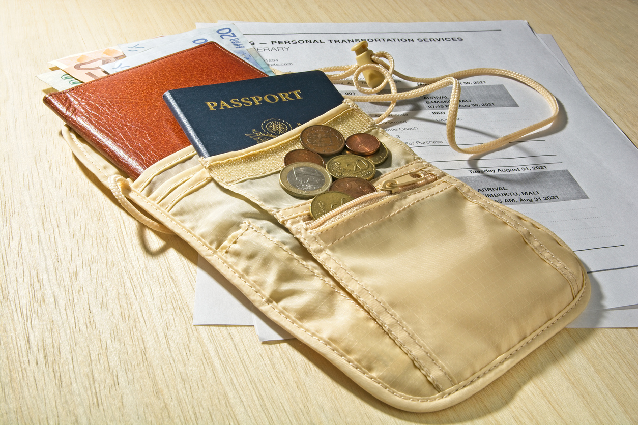 Security pouch with travel itinerary, foreign currency, passport and billfold on table. Side lighted beige pouch with American passport, brown leather bill fold with Euro paper currency and coins laid on top of printed itinerary for travel to Timbuktu Mali. Background is a light wooden tabletop