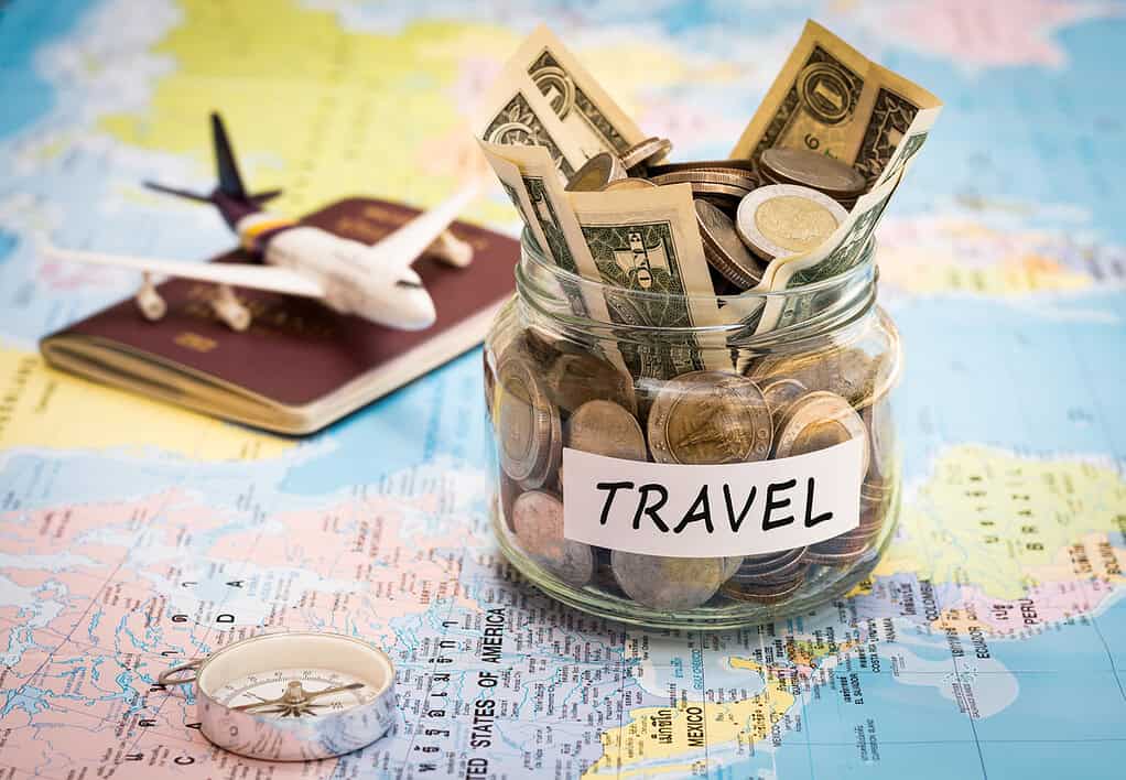 Travel budget concept. Travel money savings in a glass jar with compass, passport and aircraft toy on world map.  Budget-friendly travel plan for affordable trips